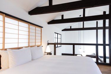 a upstairs sleeping loft has stark white walls and bedding, exposed beams, and a shoji screen