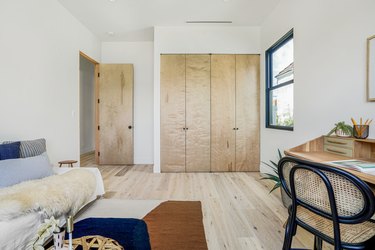 Bedroom with blue-beige-brown pillows and rug, light wood closet doors and floors, and a desk with a black frame cane chair