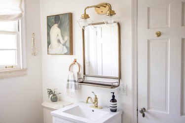 Bathroom with a figure painting, gold faucet sink, mirror with a shelf, leather and wood ring towel holder, floral accents