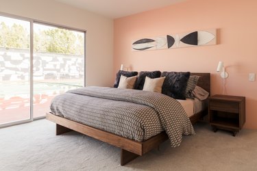 Pink wall bedroom with black and neutral bedding. A wood night stand, lamp wall sconces, and abstract painting. A large picture window.