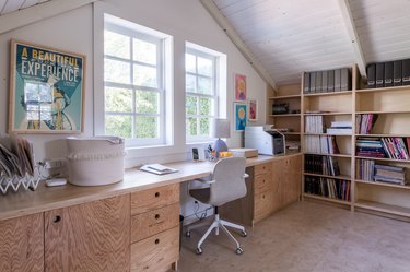 A wood Scandinavian style desk, by a window, and a gray office chair. A wood bookcase with storage files and books.