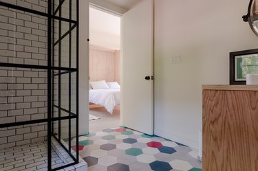 A bathroom with a red-green-blue-gray hexagon tile floor. A wall with black grout and white tile. A shower enclosed with glass and bars.