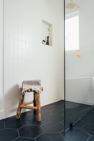 Shower with white tile, brass or gold showerhead, wood stool, and gray or slate floor tiles. Frameless glass shower.