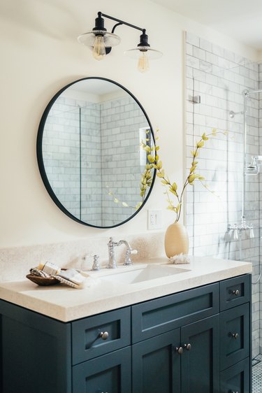 Bathroom with blue painted cabinets, white sink, round mirror, and black and glass lighting fixture. Ceramic vase on sink.