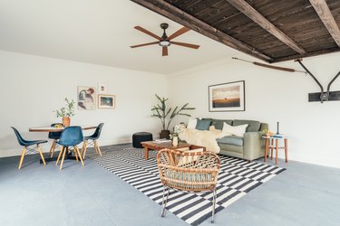 Living room in a California-bohemian style: cement floor, wood ceiling, ceiling fan, black and white rug, rattan accent chair, plant, coffee table and couch.A renovated garage with living area, cement floor, and wood ceiling.