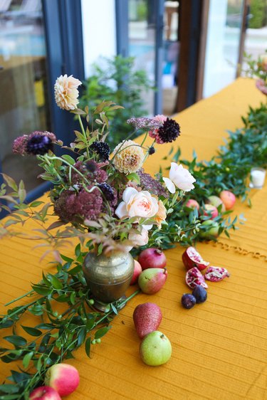 a gold-colored table with a runner made from greenery, flowers, and fruit