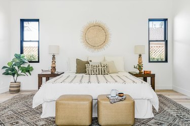 Bedroom with beige and white color scheme. A cube ottoman with teacups, a bed with beige-white bedding, woven wall hanging