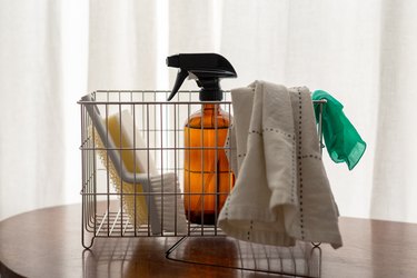 A metal basket with a washcloth, gloves, spray bottle, cleaning brush, and sponge