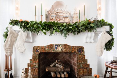 A white fireplace with a brick heart covered in a green holiday garland, two white stockings, and tapered candles.