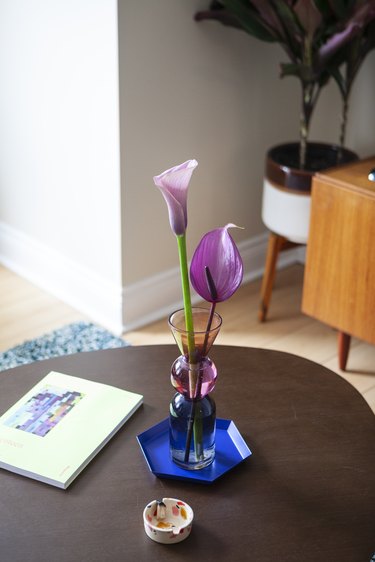 A wood table with a Harvard GSD Magazine No. 44, a pink lily in a tri-color vase with varied shapes, and a blue hexagon coaster.