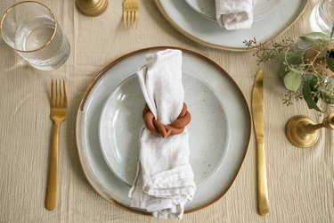 Dining table with gold flatware, white plates, cloth napkins and braided clay terra-cotta napkin rings