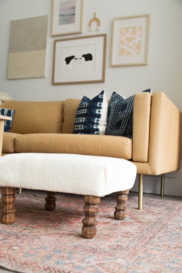 A boucle covered ottoman with wood table legs and a beige sofa with blue throw pillows and a gallery wall