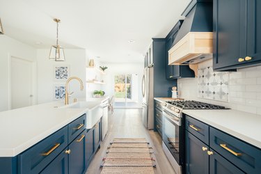 Dark blue kitchen cabinets with gold handles and white countertops. Ornate backsplash by stove top. Gold lantern pendant lights.