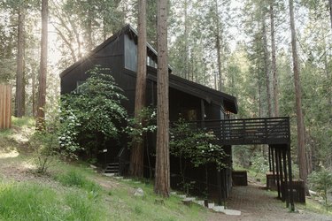 A black house with a deck, picture windows, and slanted ceiling. Forrest trees surround.
