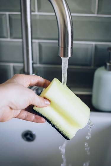 Hand holding yellow sponge under silver metallic faucet in white sink against turquoise backsplash