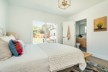 A bedroom with a picture window, gold ceiling lantern, contemporary wall art, and a bed with white bedding and colorful pillows