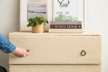 Person pulling a round handle of an IKEA Tarva dresser, covered with burlap, with plants, art, and books on top