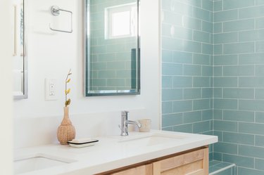 Bathroom with blue subway tile shower, vanity with white counter-wood cabinets, rectangular mirror and a vase with a dry plant cutting