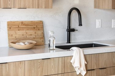 Spray bottle of cleaning liquid on kitchen counter with beige accent bowl and wood cutting board and black faucet sink.