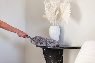 A person dusting a black half-moon side table with a vase of dried florals