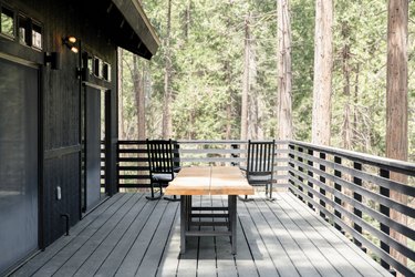 A wood picnic table and two black rocking chairs, on a deck of a black house. Forrest trees surround.