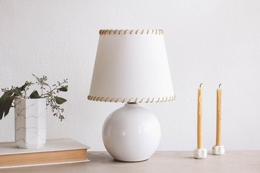 SKOTTORP lampshade with leather whipstitching, on a table with candlesticks, a book, and a vase of flowers