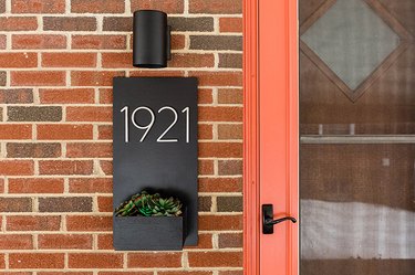 Black sconce light over a number plaque with a succulent box, attached to a brick house, with an orange door