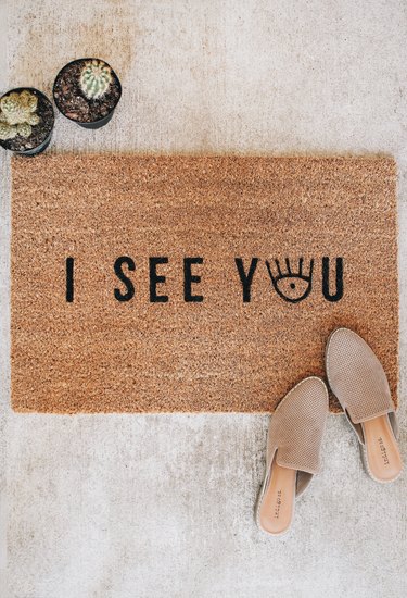 a woman's shoes on a brown doormat with black letter spelling "out i see you"--the "o" in "you" is an eye graphic