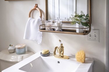 Bathroom sink with a gold faucet, mirror with an attached shelf, leather and wood ring towel holder, and floral accents