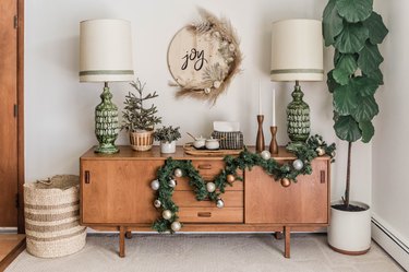 Round wood sign with 'joy' written in black cursive and half moon dried florals, over a wood sideboard with Christmas decor