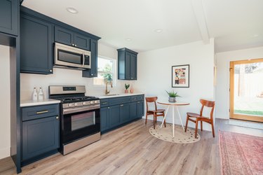 Kitchen with dark cabinets and small dining room corner with wood chairs and a plant on a white round table on light wood flooring