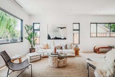 Living room with a round fiber rug, glass and stone coffee table, white sofa with multicolored pillows, windows of varied sizes, and a palm