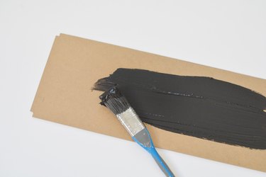 Blue handled paintbrush applying black paint to a rectangular cardboard panel against a white background