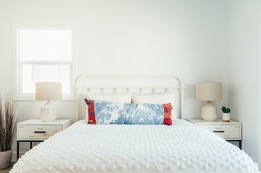 A bed with white bedding and colorful pillows, a white nightstand and lamp, and houseplants