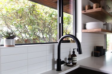 A black sink faucet, by a window in a kitchen with a white backsplash and wood shelves.