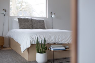 Neutral wooden platform bed under window with 2 reading lights on either side and table and plant at foot of bed