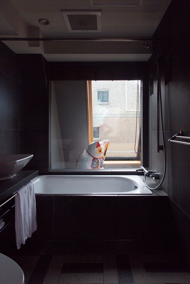 a deep white tub contrasts with dark gray walls and floor; a waving lucky cat figurine is visible through the bathroom window