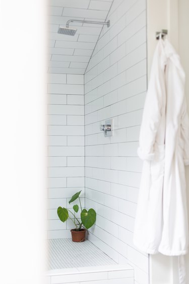 white subway tile shower, silver overhead showerhead, potted plant in the corner of the shower, white bathrobe hung up