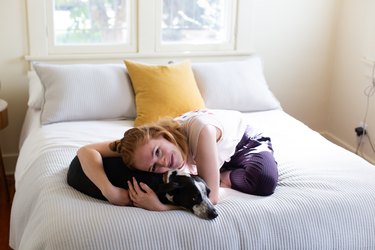 a woman hugs her dog on a bed with a striped duvet and one bright yellow accent pillow