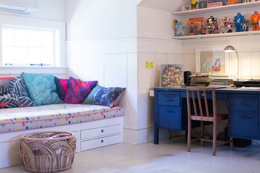 Kid's room with a blue desk, action figure toys, tie dye pillows, window seat with polka dot cushion, and Boho basket.