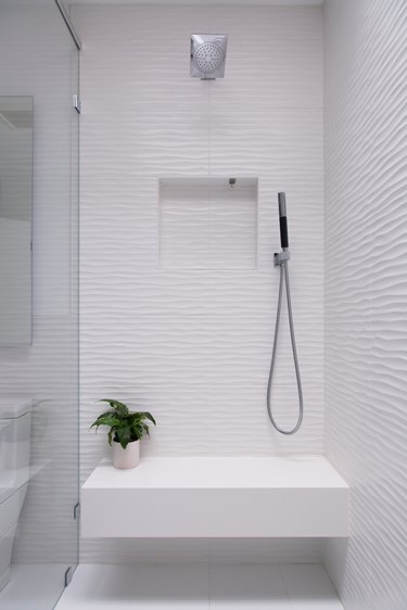 white shower with bench that has a small plant on it, glass door, overhead showerhead and handheld showerhead