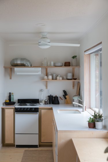 Kitchen corner with stove and open shelving above with dishes and glassware knife, cutting board and coffee pot below kitchen sink under window