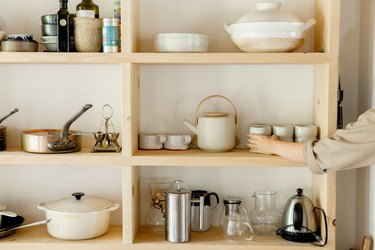 Wood shelving with white and beige dish ware, coffee pots, and pans. A person is reaching for a mug.
