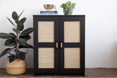 Black IKEA Brimnes cabinet with added cane doors, accented with plants, books, and a gold bowl; rubber tree plant