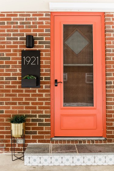 Front entrance of a house with an orange door, black sconce light, gold plant stand, brick exterior