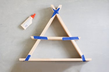 Wooden a-frame shelf taped together along edges with blue painters tape next to glue bottle against grey background