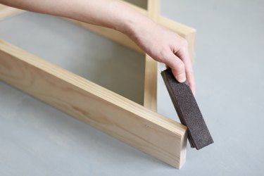 Hand using sanding block to sand edge of wooden a-frame shelf against grey background