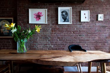 A natural wood table with a vase of yellow flowers. A brick wall has eclectic art.