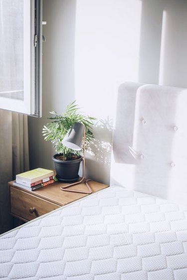 White mattress next to side table with lamp and plant in white-walled room with white curtains