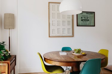 Dining room table with plastic chairs in white-walled room with small wood cabinet, framed graphic artwork, and lamp in corner